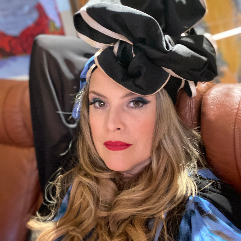 A woman with fair skin and long wavy blond hair sits on a brown leather couch, one arm relaxing behind her head, she is looking at the camera. She has bright red lipstick and smokey eyes, she wears a large ruffled black and white headpiece.