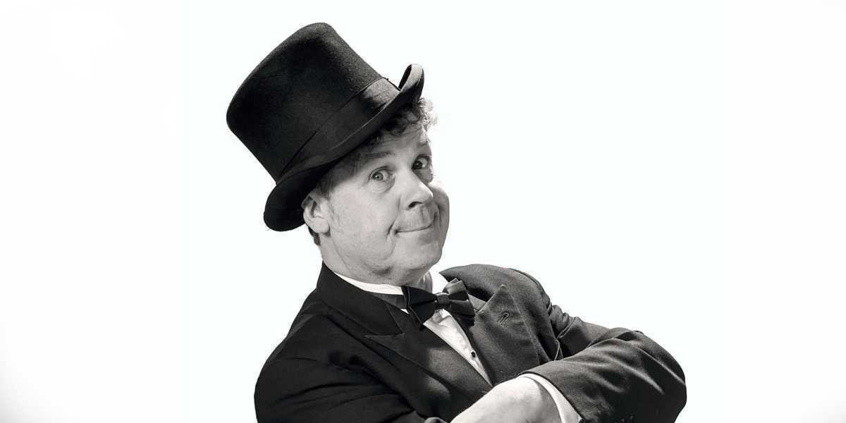 Raymond Crowe facing the camera. He is wearing black suit, white shirt and a black top hat standing on a white background.