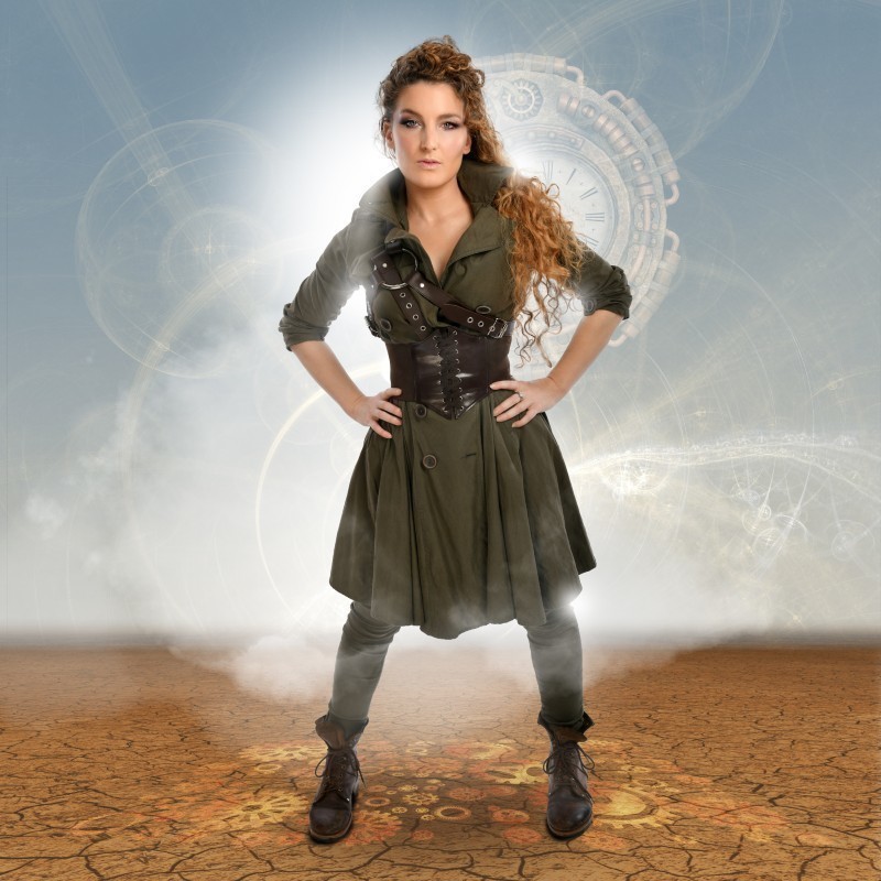 Alice Fraser: Chronos - Alice Fraser stands with her hands on her hips. She wears a green long coat with a black corset and detailing over the top of the coat. The background is light blue with a transparent image of a watch behind her.