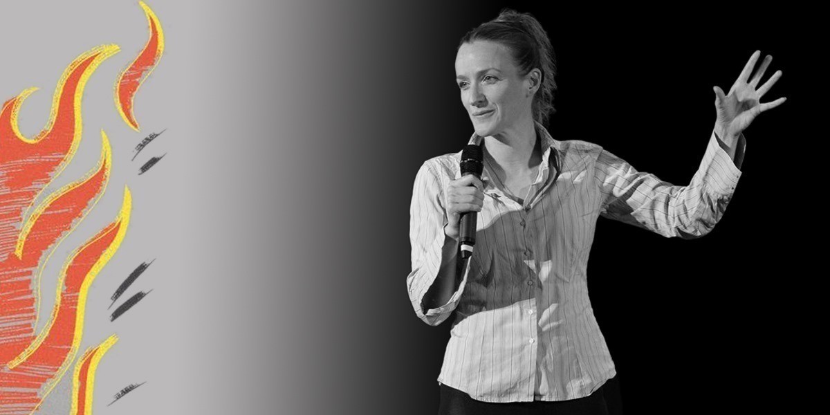 Kate Smurthwaite holding a microphone and raising her other hand in black and white with hand-drawn flames to the left of the image.