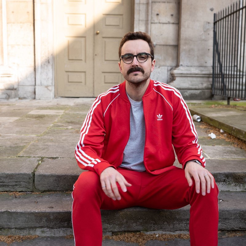 A photo of a man sitting on a step wearing a red tracksuit with white stripes and a grey t-shirt with a blank expression on his face. He is wearing black framed glasses and has a prominent brown moustache.