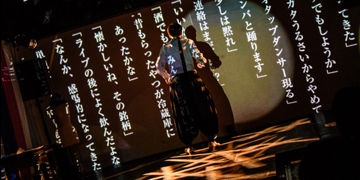 Japanese characters projected on a screeen with a man in the centre with his back to us.