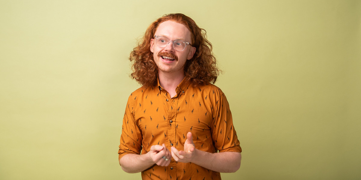 Nick stands in front of a light green backdrop with his long curly red hair out. He's staring off to the right, hands gesturing and mouth open as if he's mid story.