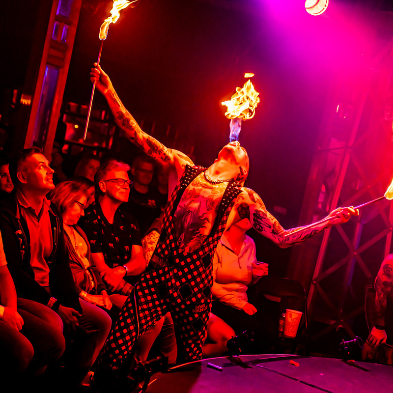 A tattooed man in polka dot overalls breathes fire surrounded by an audience.