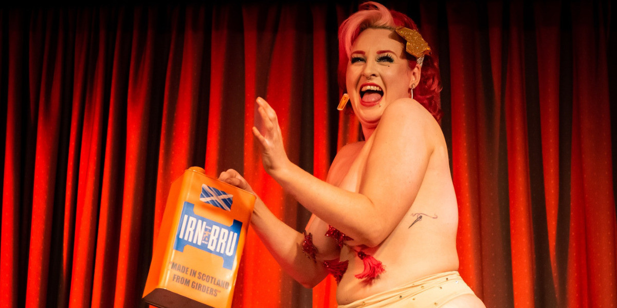 Roxy Stardust stand on stage in front of a red curtain, she is smiling wide. In her right hand she holds a Jerry can of Irn Bru.