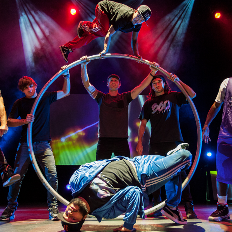 A group shot of the ALLSTARS featuring a BMX rider, basketballer, breakdancers, acrobat and musicians, facing the camera under colourful stage lighting.