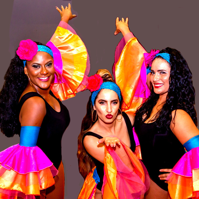 A photo of three people posing. They are wearing black bodysuits with pink and orange frilly sleeves on their arms, as well as blue headbands adorned with a pink flower. The person in the middle of the photo is blowing a kiss to the camera and the other to people on either side have one hand placed on the wall behind them.