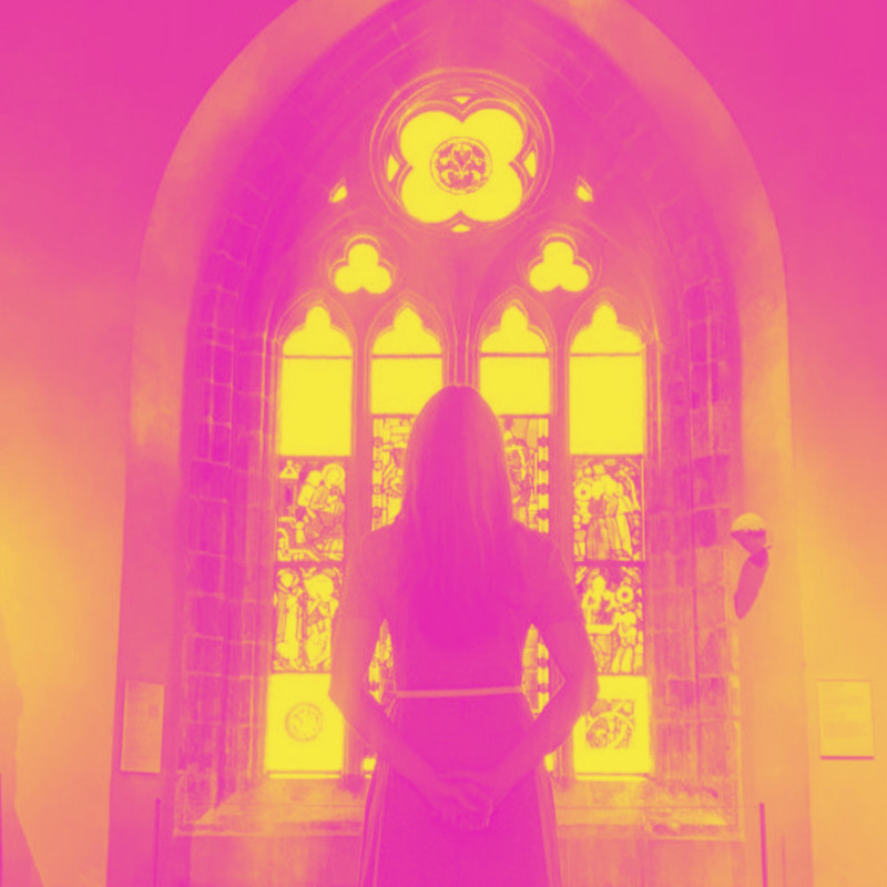 Child of Sunday - The artist stands silhouetted, facing a stained glass window of an old church, hands clasped behind her back. The image is washed in pop shades of candy pink and bright yellow.