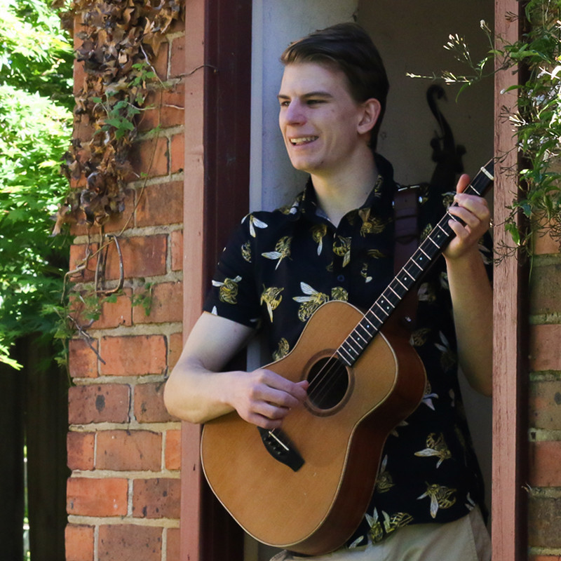 A smiling man stands in a brick doorway holding a guitar facing the left. There is ivy across the brick walls and trees in the distance to the left of the building. The man has short brown hair and wears a black short sleeved shirt with large pictures of bees all over it.