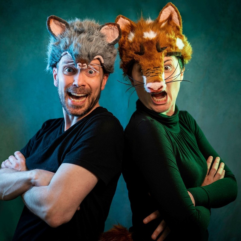 Two performers standing back to back with heads slightly turned facing the camera. One performer wearing a fox costume for her head and other performer wearing a snake one. They are making screaming faces.