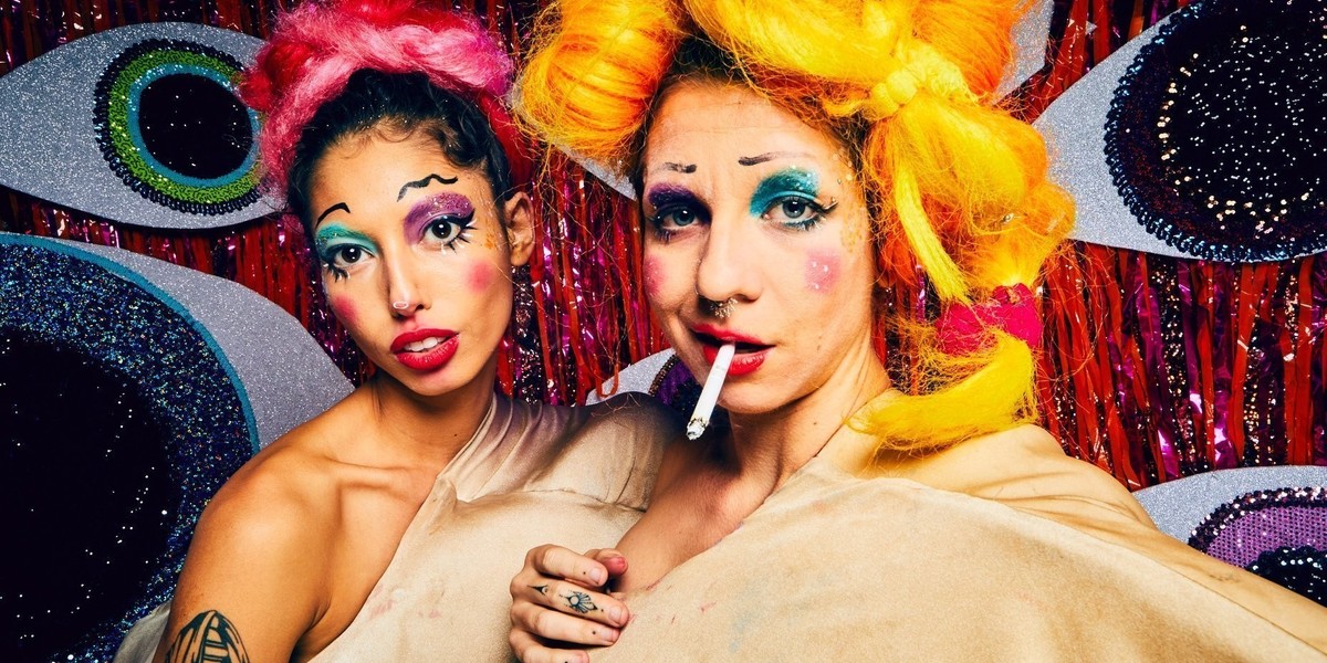 Two clown showgirls stair at the camera. One of them is smoking a ciggeret.