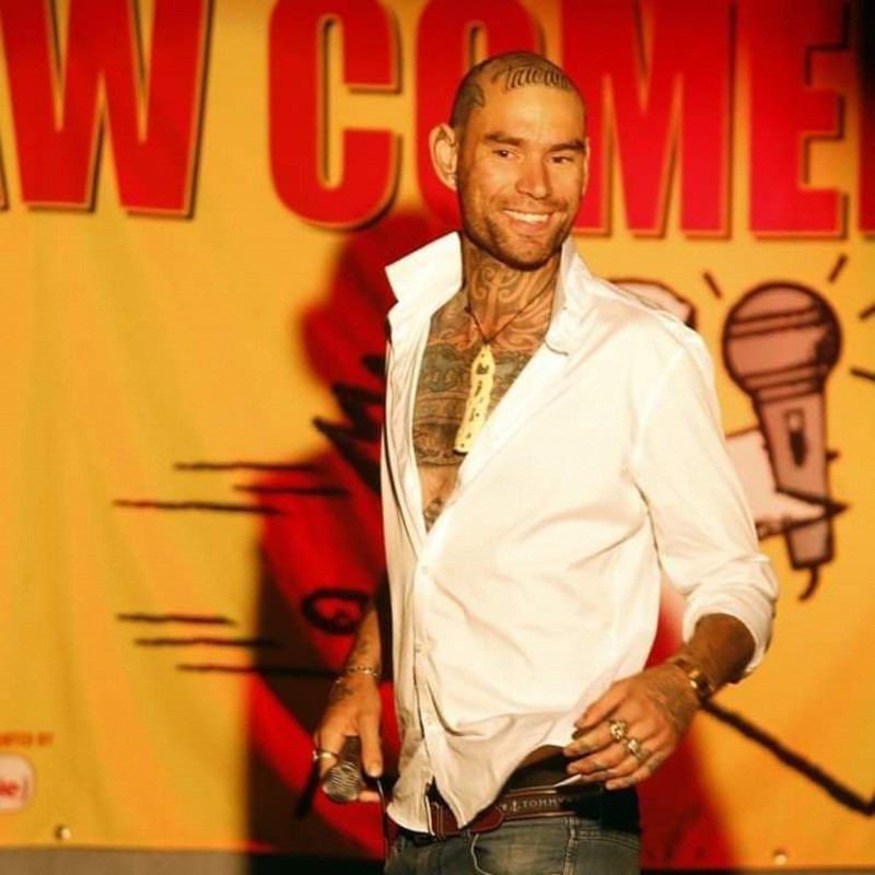 Rudes gives head... Liners - A photo of  a guying wearing a white shirt smiling. He is wearing a necklace with a large cream coloured pendant. The top buttons of his shirt are undone, with his tattoos visible across his chest, on his forearms on on his head.