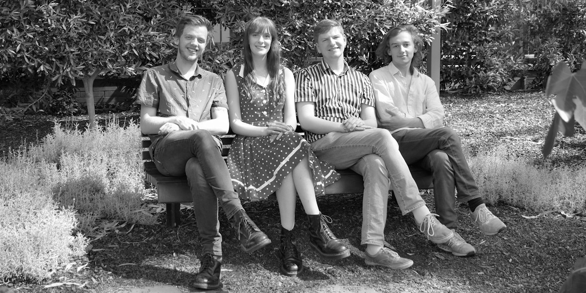 Four performers (Left to Right: Jack Barton, Tiffany Gaze, Dan Courts and Edmund Black) sitting on a bench in a garden setting, all with their legs crossed in the same direction.