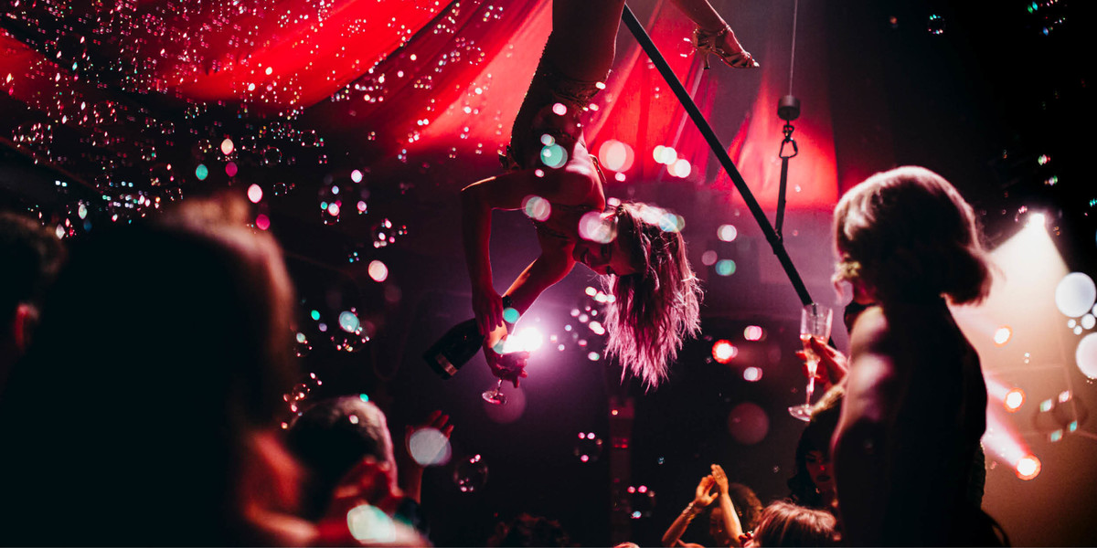 A beautiful performer in a gold bodysuit swings above the audience in a room filled with bubbles serving a glass of champagne in Blanc de Blanc Encore