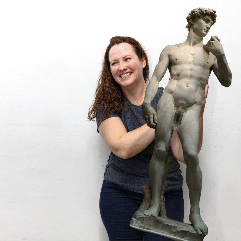 Archaeologist / comedian, KC Martin-Stone, looks off camera with a cheeky smile on her face. She's holding. photoshopped statue of Michaelangelo's David, whose genital area is pixelated. KC wears jeans and a grey t-shirt, and stands against a white background.