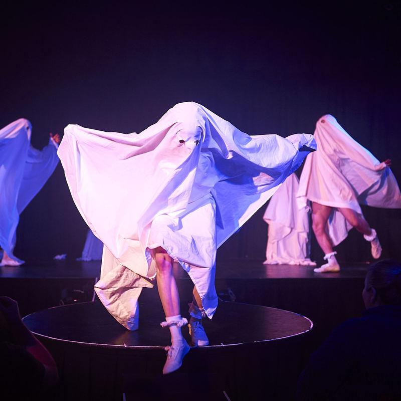 Three performers running off stage in different directions dressed as ghosts with white sheets over their heads.