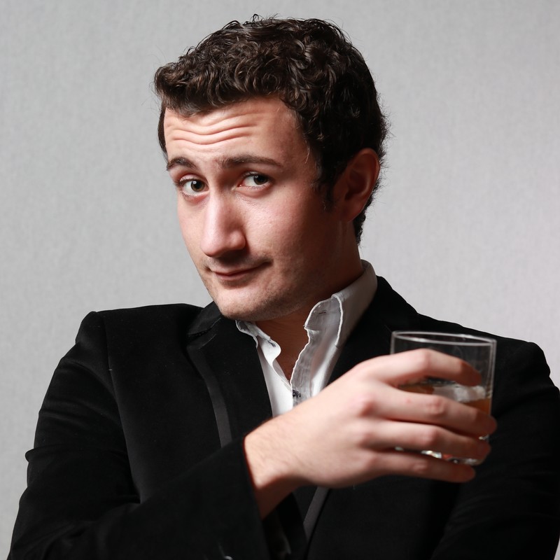 A photograph of a man wearing a white shirt and black suit jacket. He is holding a glass of water across his body. He has short brown hair, while smirking at the camera.