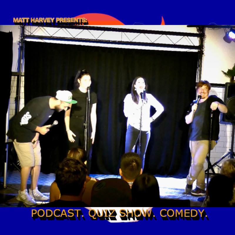 A photo of four people on stage talking and laughing into microphones. Behind the stage is a black curtain. The text on the background on the photo reads, ‘Matt Harvey Presents’ ‘Podcast. Quiz Show. Comedy’ in yellow capital letters.