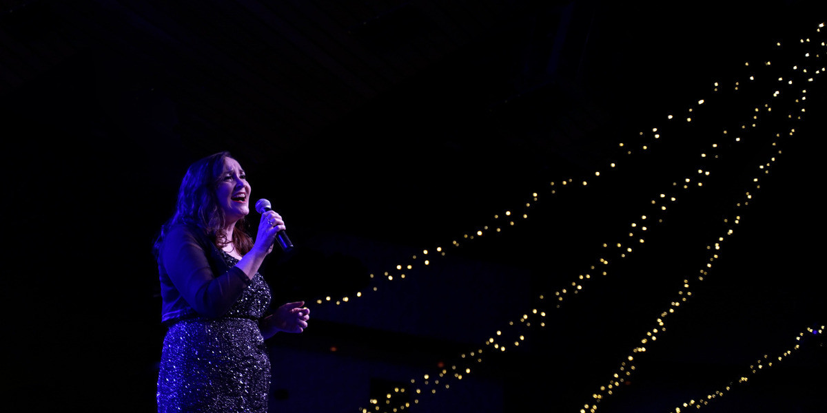Deborah Caddy singing into a microphone underneathe strings of fairy lights.