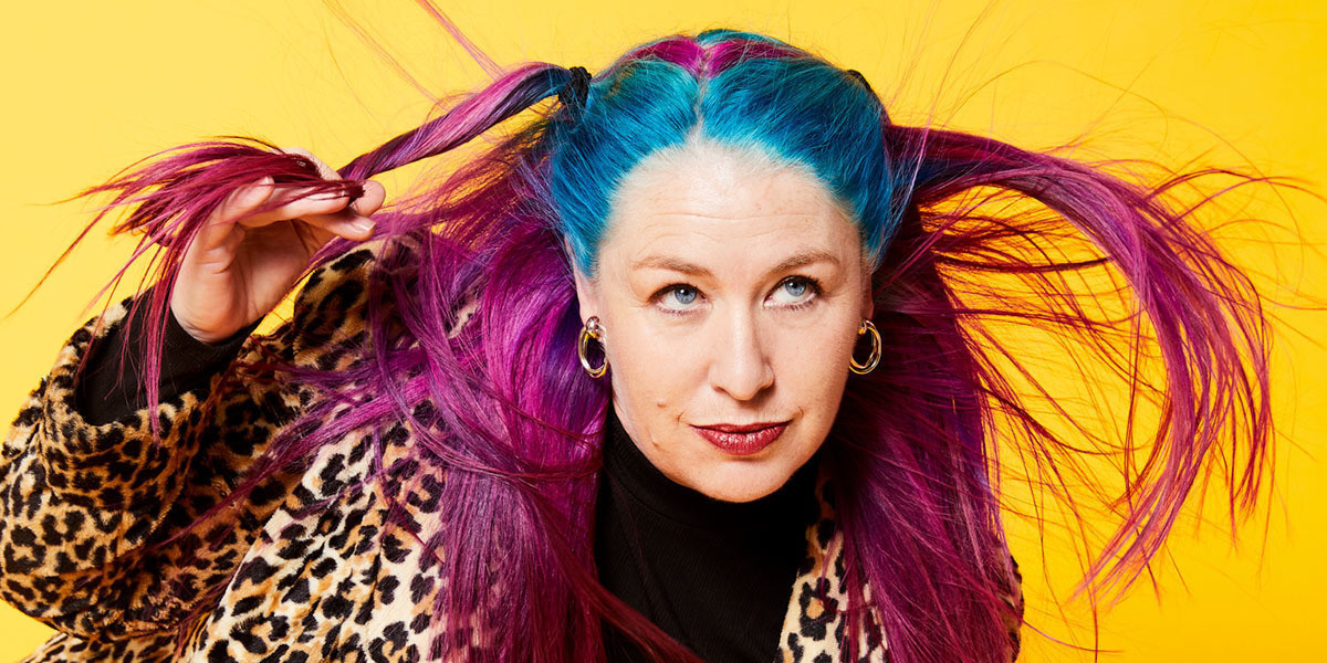 A close-up of Kelly Mac against a yellow background. She twirls a strand of her long pink and blue hair around a finger. She is Caucasian and looks off camera.