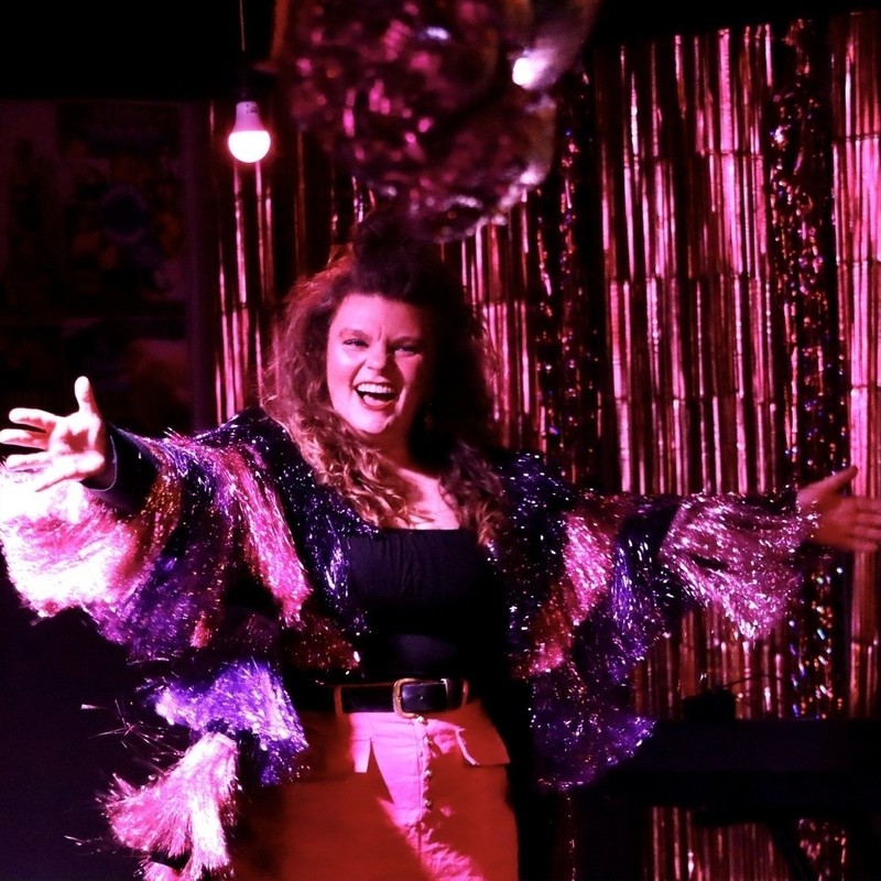 The Finest Filth Variety Hour - Annie one of the hosts of The Finest Filth Variety Hour smiles on stage as she extends her arms dancing. She has long curly brown hair and is wearing a pink and purple tinsel jacket with a pink skirt.