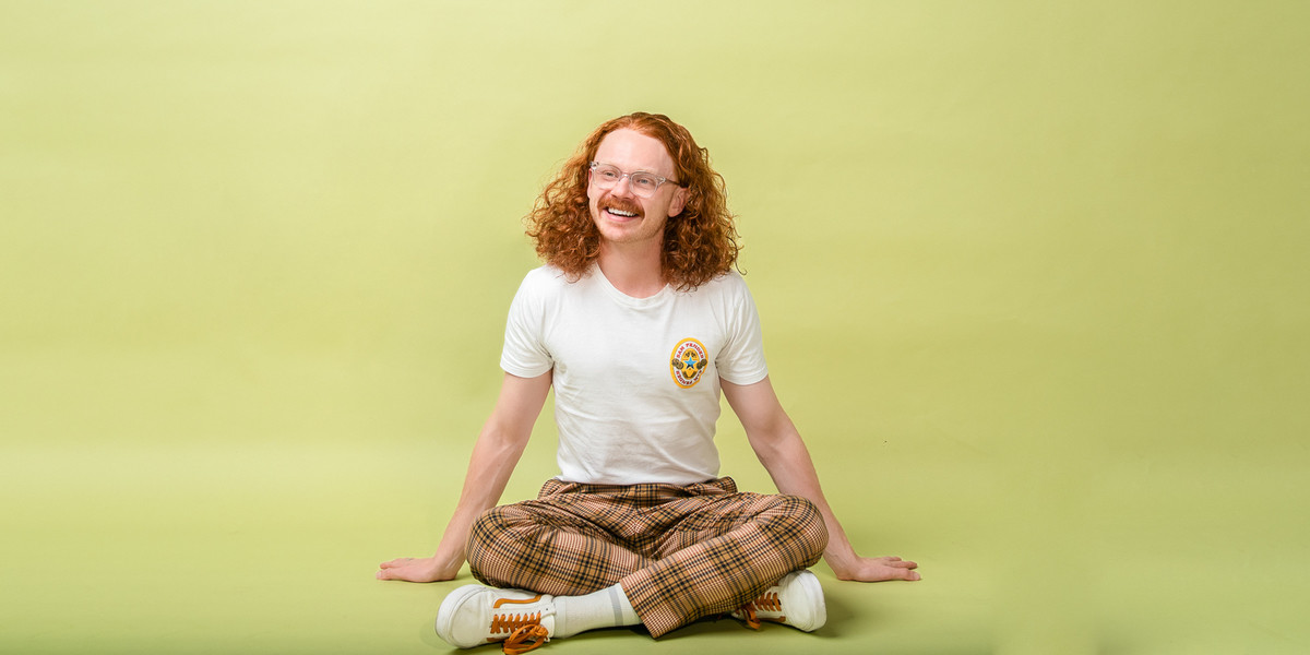 Nick sits crossed legged in the centre of a light green backdrop with his long curly red hair out. He's wearing orange and brown checked pants and a white tee shirt. He's got a gleeful smile staring off to the right.