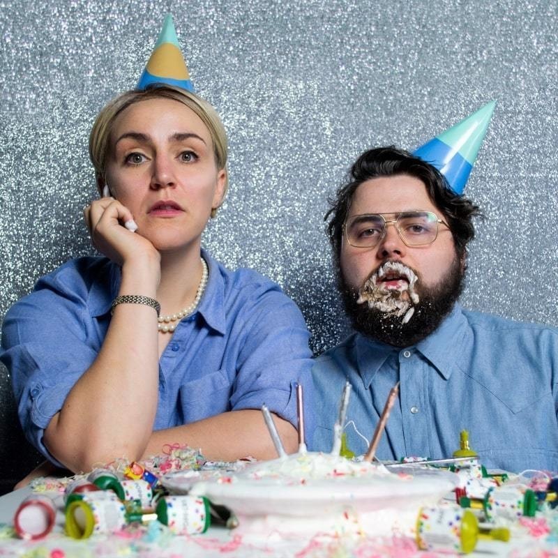 A man and a woman sitting in front of a bright gray background. The two wear a blue long-sleeved shirt and party hats. There's a birthday cake with candles in front of them and decorations around it. The man has a brown beard full of icing from the cake.
