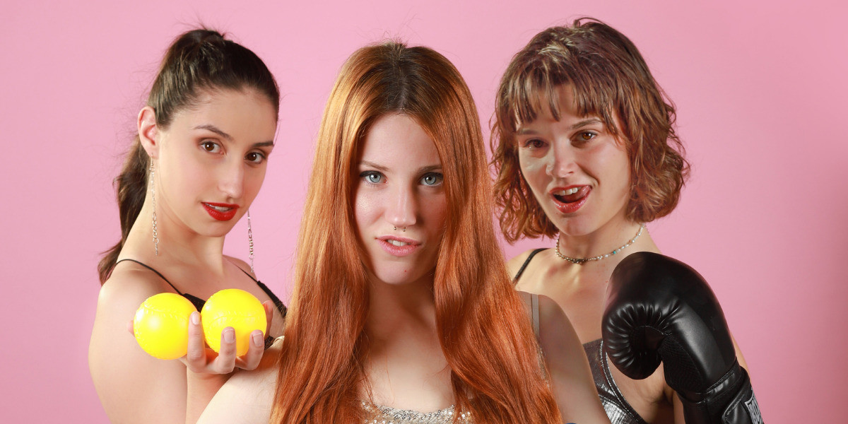 Hit n Hope: A Character Cabaret - Three women posing in front of a solid pink background. Steph (left) has dark hair in a ponytail, wears long silver earrings and holds two yellow balls in her hand. Tess (centre) has long red hair and stares with a pouty smirk. Nic (right) has mid length brown curly hair with a fringe, and wears a pearl necklace, is smirking with her tongue out, and is wearing a black boxing glove on her hand.