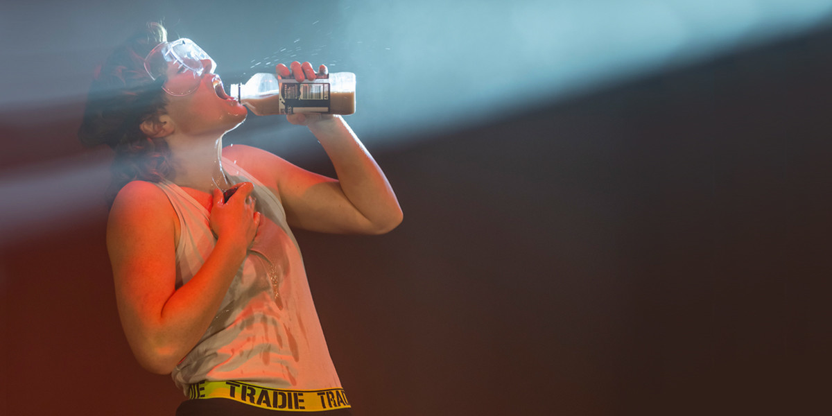 A performer from DEADSET holds a chocolate milk bottle to her lips, while standing in a spotlight. She is wearing safety goggles, a white singlet, and Tradie branded underwear. She stands profile into the spotlight, and sips the chocolate milk, with the drink spilt down the front of her singlet
