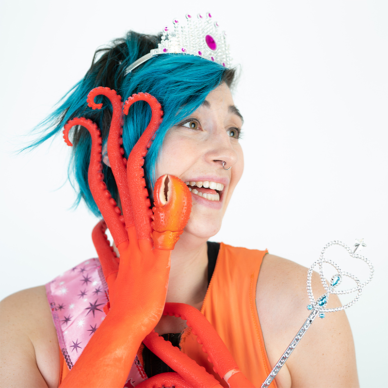 A transgender person with blue hair and squid tenticles for fingers, wears a plastic princess tiara and clutches a sparkly septer, smiling proudly.