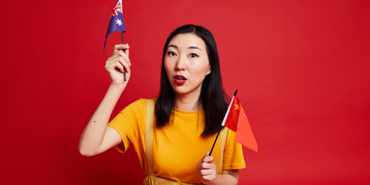 Jenny Tian - Chinese Australian: A Tale Of Internet Fame - Jenny is looking directly at the camera with her mouth gaped. She is holding an Australian flag in one hand and a Chinese flag in the other. She is wearing a yellow top and overalls and sitting infront of a red backdrop.