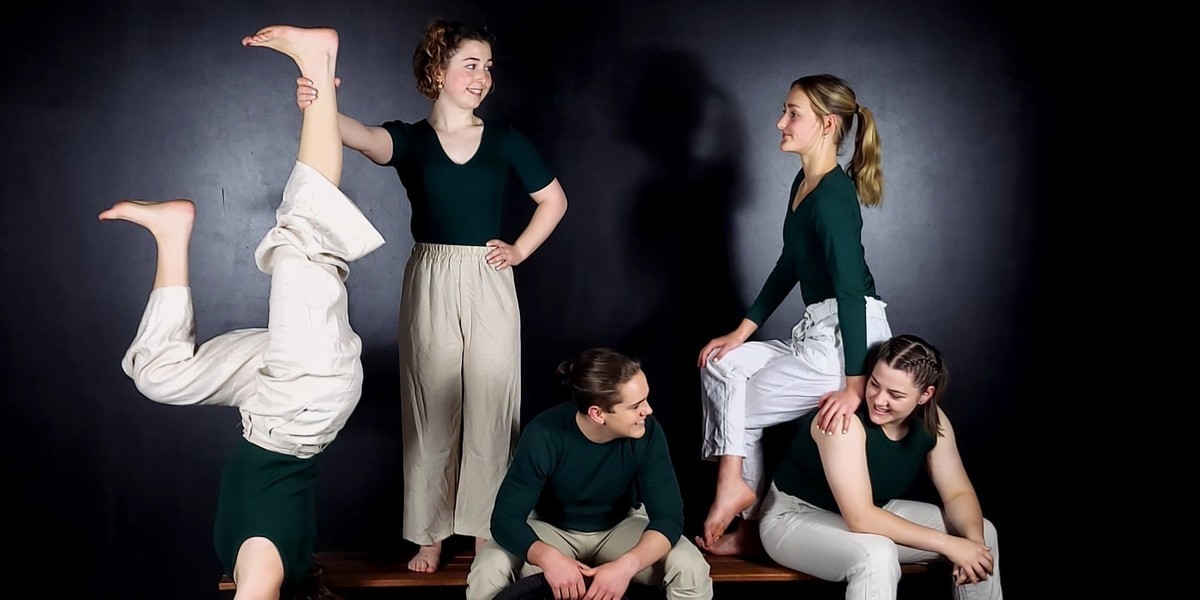 5 teenagers positioned on a bench wearing cream pants and dark green tops with a black background. 1 teenagers is holding a unicycle and another is in a handstand.