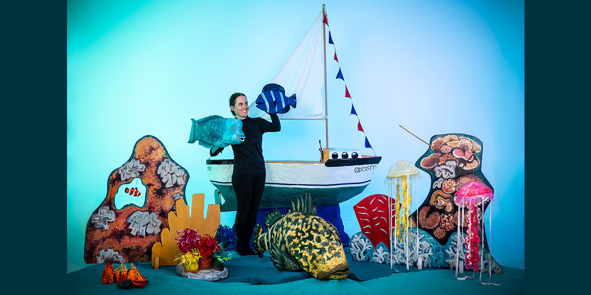The puppeteer stands in front of a sailing boat holding two fish puppets.
