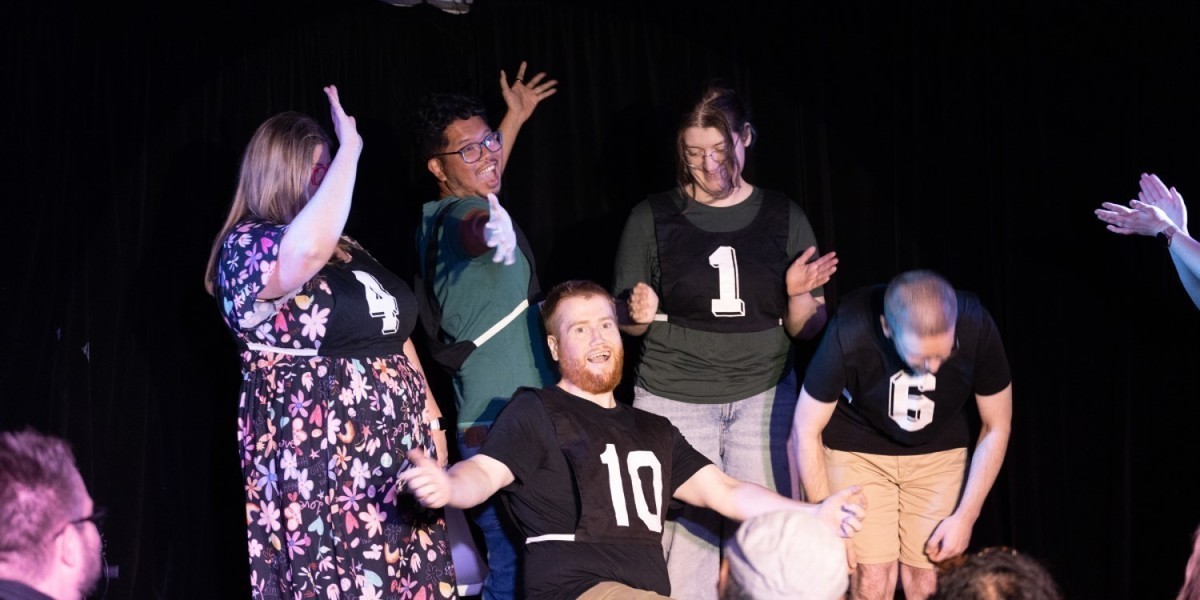 A group of five improvisers on stage wearing netball bibs. Each bib has a number on it. The improvisers are arranged as if they have just finished performing a song. One improvisers is bowing and others are holding up their hands with a flourish.