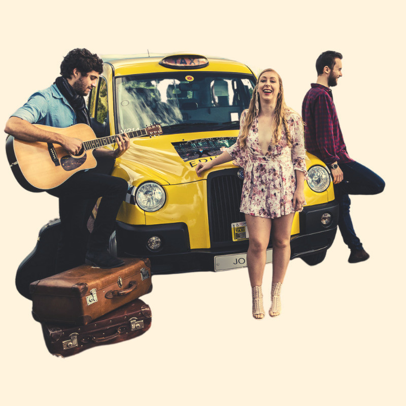 A woman with long golden curls stands in front of a yellow taxi. She is wearing a pink floral playsuit with feathers in her hair and her head is tilted back as she appears to be laughing. To the left of her is a man in a blue shirt and black scarf. He is playing a guitar with his foot resting on two vintage suitcases on the ground in front of him. To the right another man in a red plaid shirt leans against the taxi looking in a different direction. He is also laughing.
