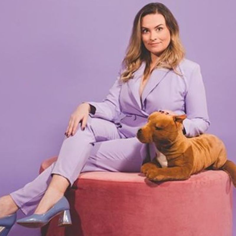 A brown haired woman in a lavender power suit and matching shoes sits on a pink velvet lounge. She is petting a large, stuffed toy dog.
