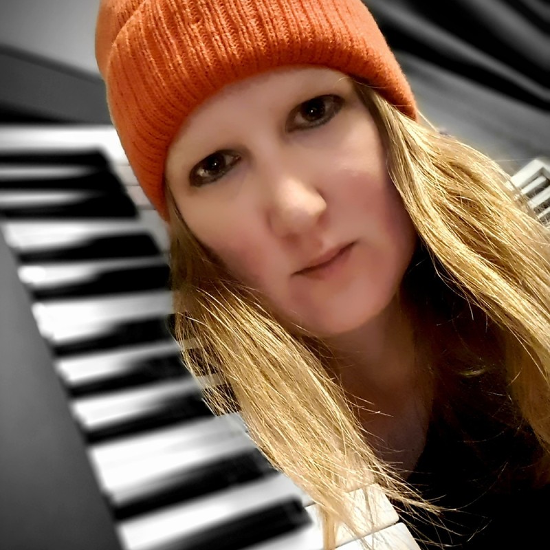 CANCELLED - Folding Minchin - Emma Knights wearing an orange Beanie and heavy black eyeliner with a piano.