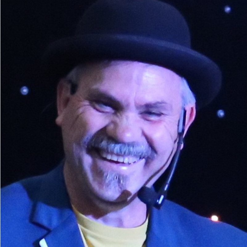 Magic on the Edge - Success - A headshot of a man smiling. He is wearing a black hat and has a earpiece microphone that sits on the side of his face. He is wearing a blue jacket and yellow t-shirt.
