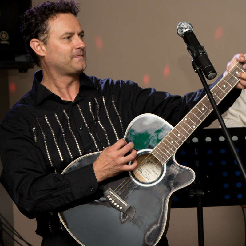 A person with short brown hair wears a black shirt with sequined stripes down the front. He plays a black acoustic guitar on a stage.