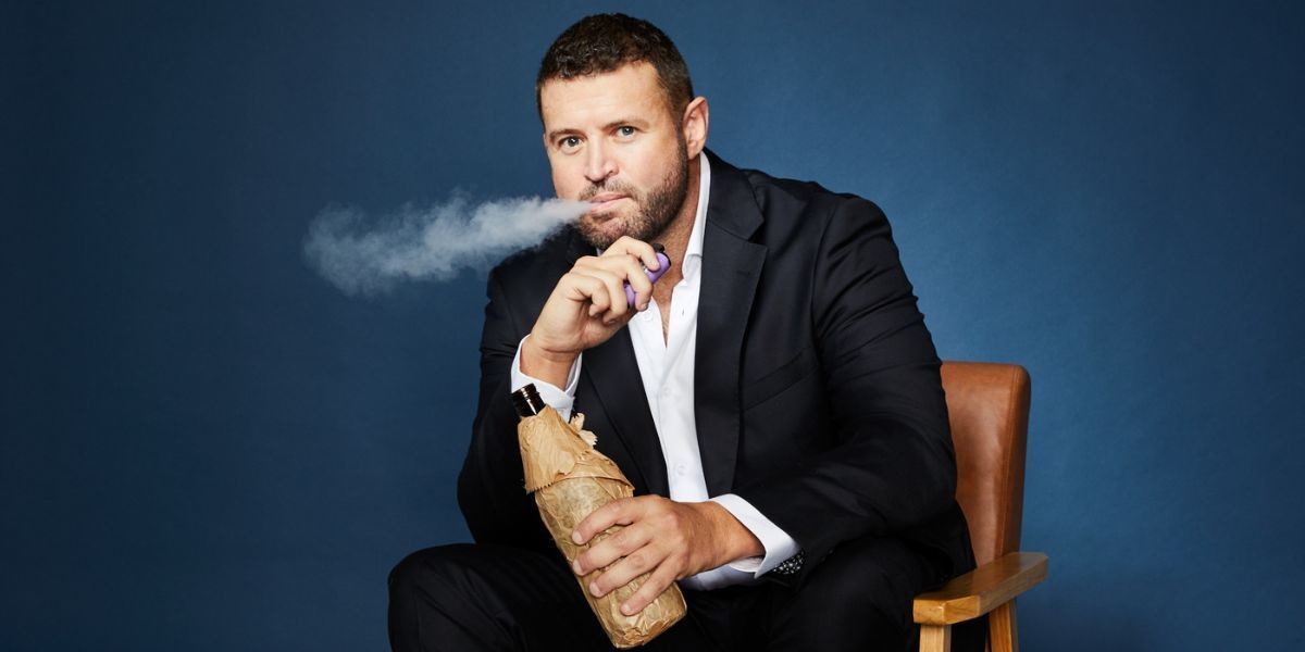 Andrew Hamilton: Sh*t Bloke - Andrew sits in a chair in front of a blue background. He holds a bottle of liquor in a brown paper bag and a vape. He's blowing out smoke.