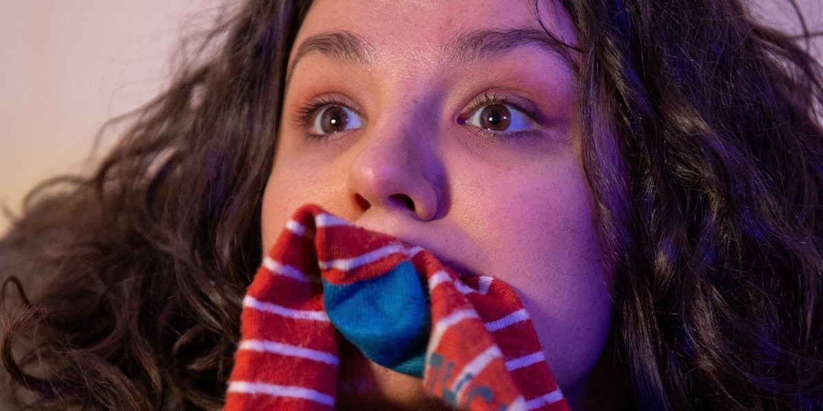 A close up photo of a woman with a red and white striped sock in her mouth. With her brown eyes wide open.