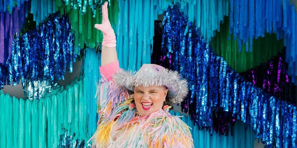 CANCELLED - SHOW PONY - Annie Schofield is posing in front of a backdrop of blue and green shimmery streamers. They are wearing a silver Cowgirl hat and a pastel pink tinsel jacket. They are have a big smile on their face and are pointing their pink gloved hand into the air.