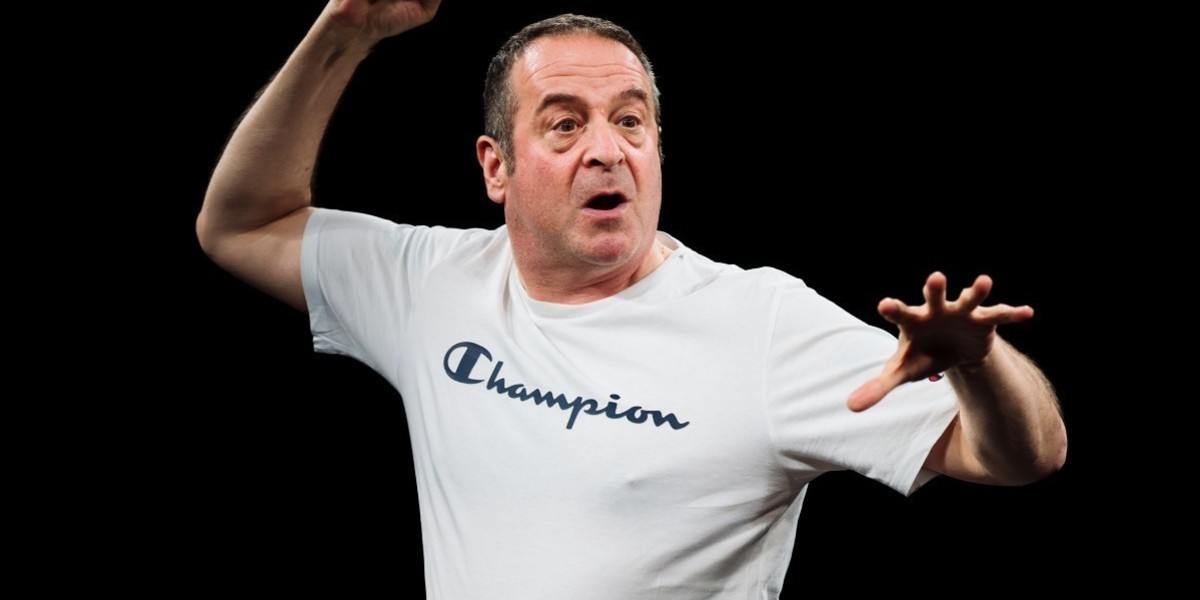 Mark Thomas in character on stage with his right arm up high and his left arm pointing forward, his mouth is open and his expression is alert and surprised. He is wearing a white shirt with black writing on it that says "Champion"