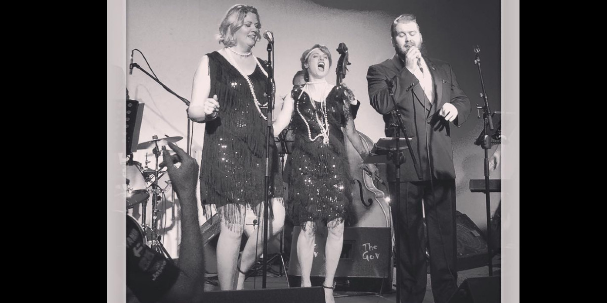 a black and white image of three singers on stage. The two women are singing and dancing and wearing vintage flapper dresses with diamond and pearl accessories. The man is wearing a tuxedo and is singing