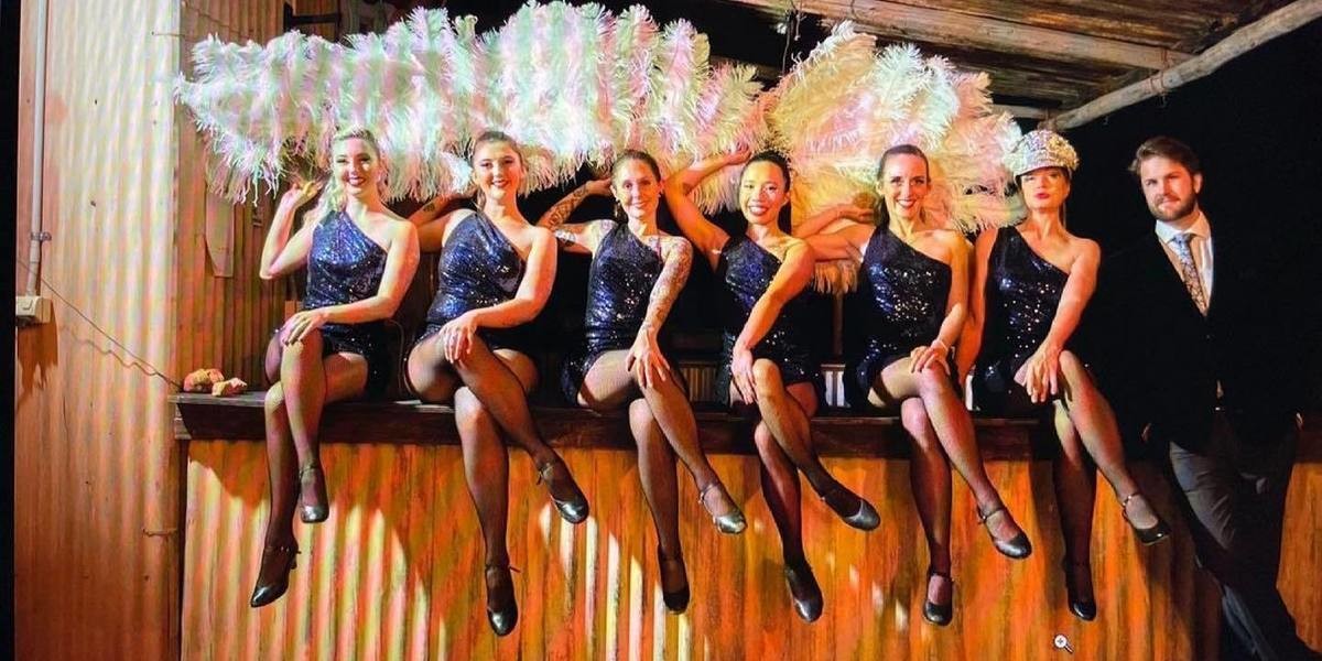 A Cabaret Cast in Blue Sequin dresses hold feather fans and pose sitting on a rustic bench
