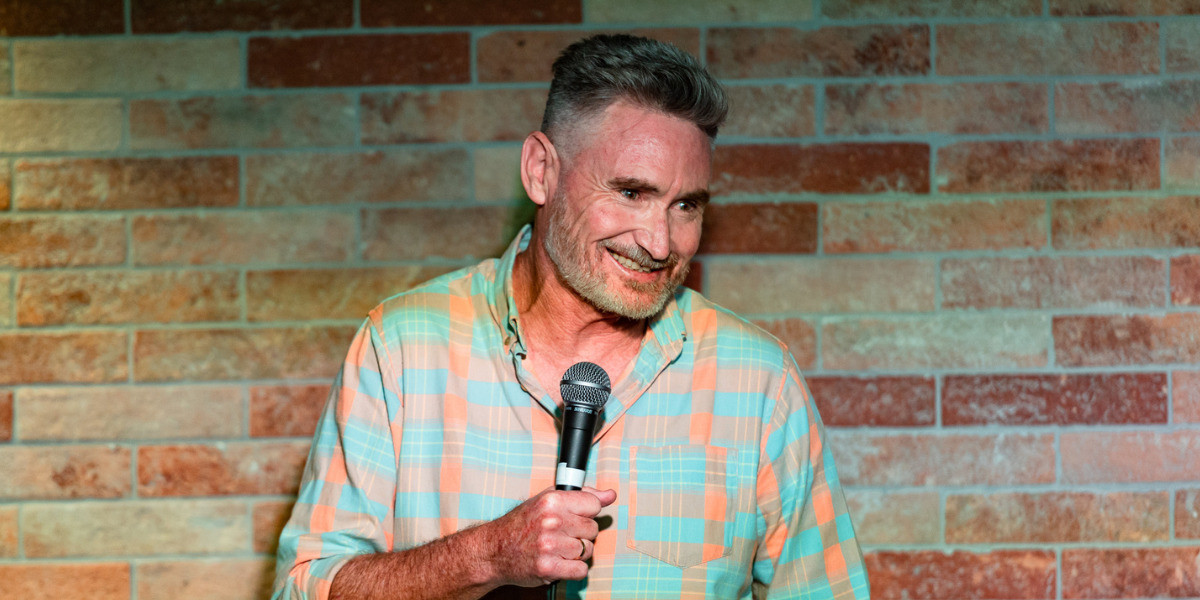 Dave Hughes - Fully Furnished - Dave is wearing a green and orange checked shirt. He is smiling while holding a microphone and looking out toward the audience. He has short grey hair and stubble. He is standing in front of a brick wall.