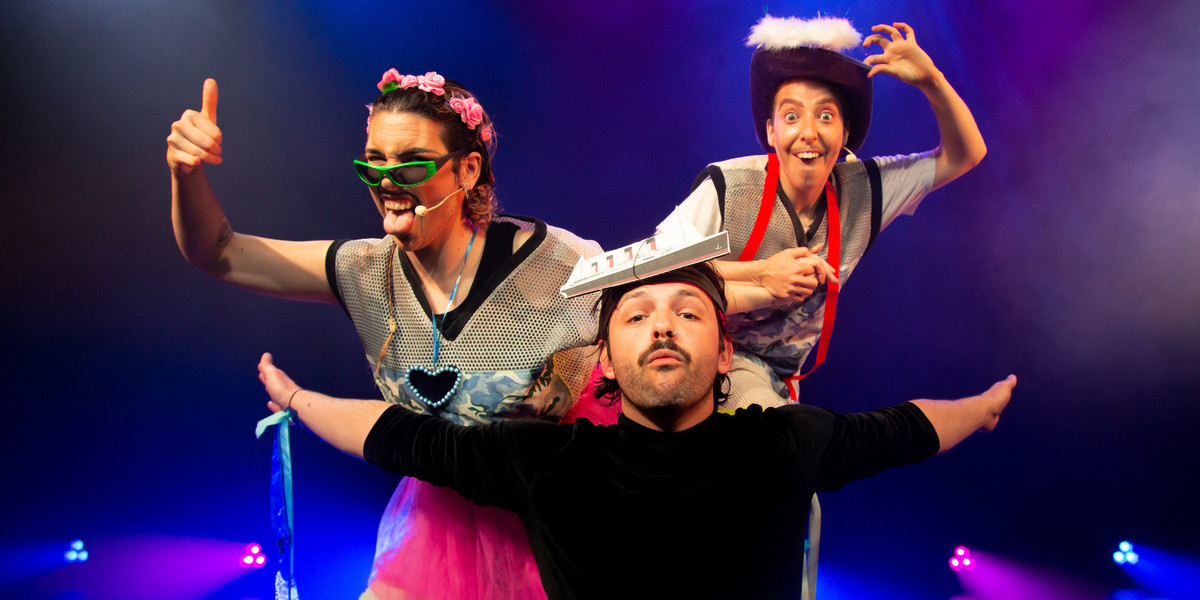 Three people are posing together on stage, the front person is kneeling with a small picture of a ship stuck to their forehead; they look serious. The middle person is leaning to the left with their tongue sticking out, wearing sunglasses and a tutu. The third person at the back is standing on a small step ladder wearing a cowboy hat and looking excited.