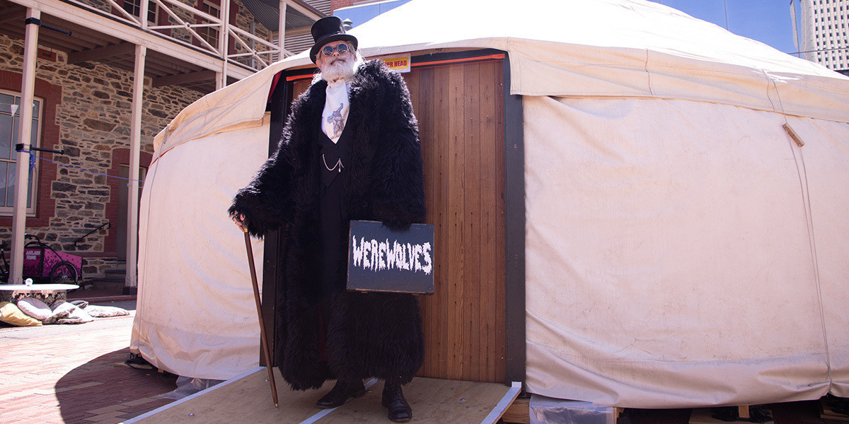 Werewolves - A gentleman stands in front of the doors of a yurt set in the courtyard of the Migration Museum, ready to perform the show Werewolves. He is dressed all in black with a very large black fur coat, top hat, walking cane and a case with 'Werewolves' written on it.