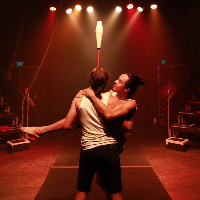 Ben is holding Simon in his arms while balancing a juggling club on his nose. There are orange and red lights in the background. They are wearing white tshirts and black shorts.