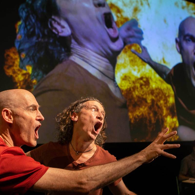 Image of two men looking into a camera yelling while their image is projected onto a screen and fire is behind them on the screen